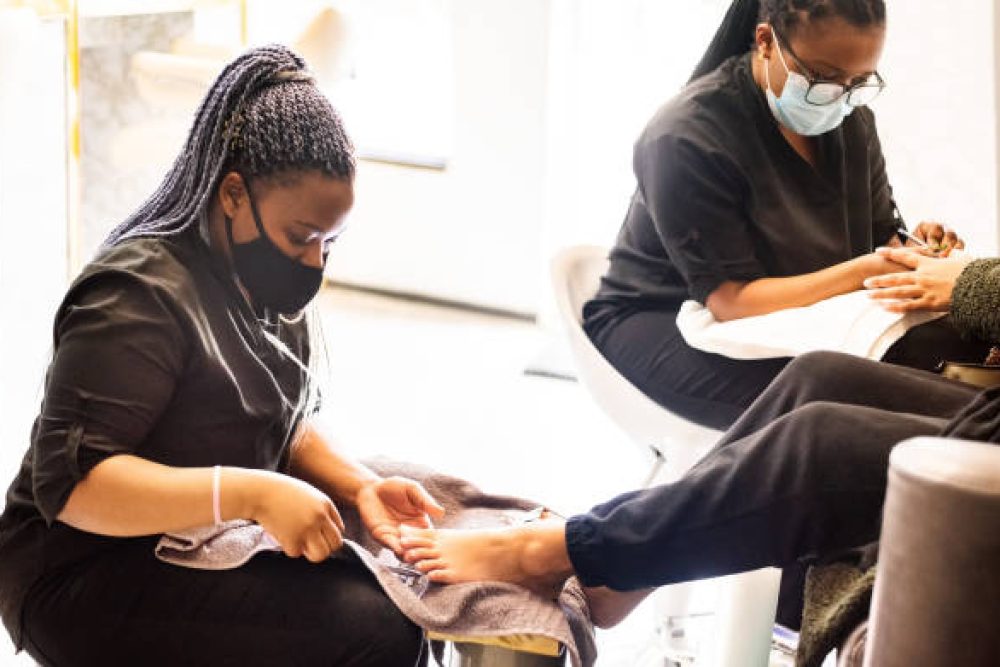 Two beauticians wearing face masks giving pedicure and manicure to a client in beauty salon